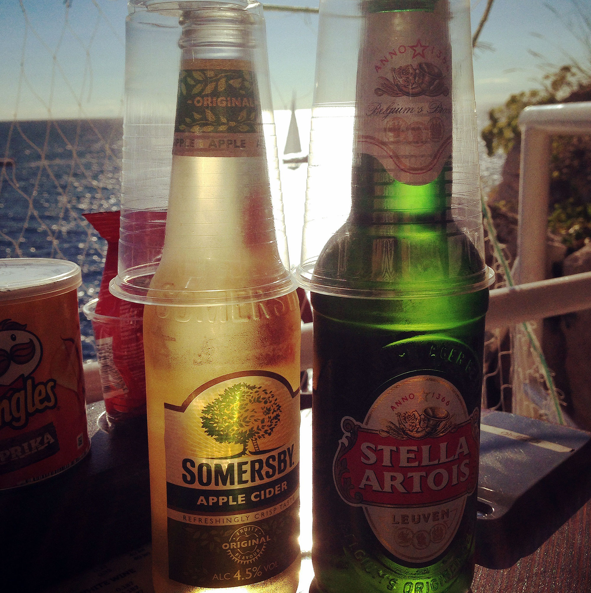 A Somersby Apple Cider bottle and a Stella Artois beer bottle sit on a table overlooking a sunlit ocean, with disposable cups placed upside down on top of each bottle. A can of paprika-flavored Pringles is partially visible to the left.