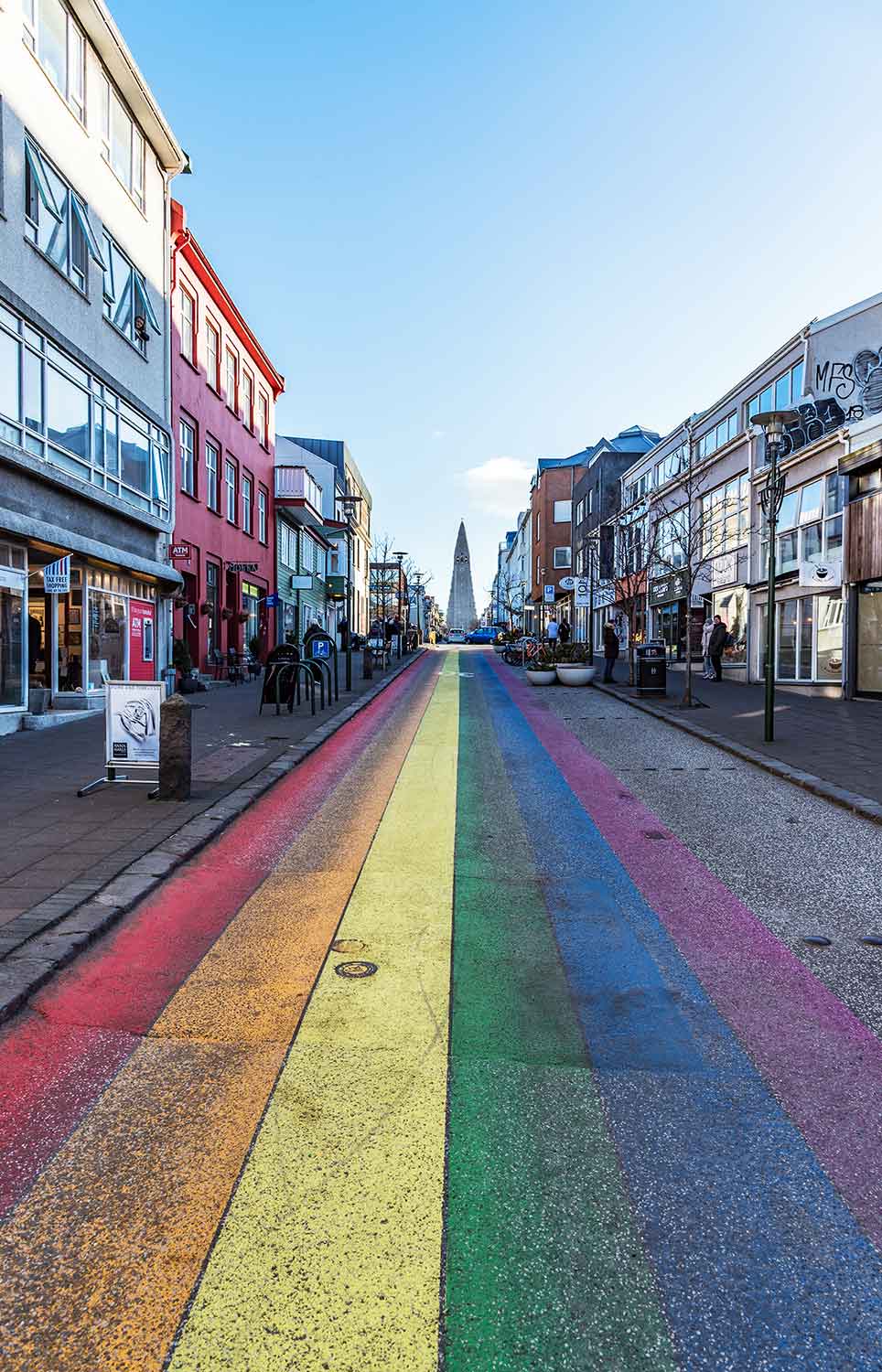 A vibrant rainbow-painted street stretches through a colorful urban area, lined with shops and buildings on both sides. In the distance, the iconic Hallgrímskirkja church towers over the scene under a clear blue sky.