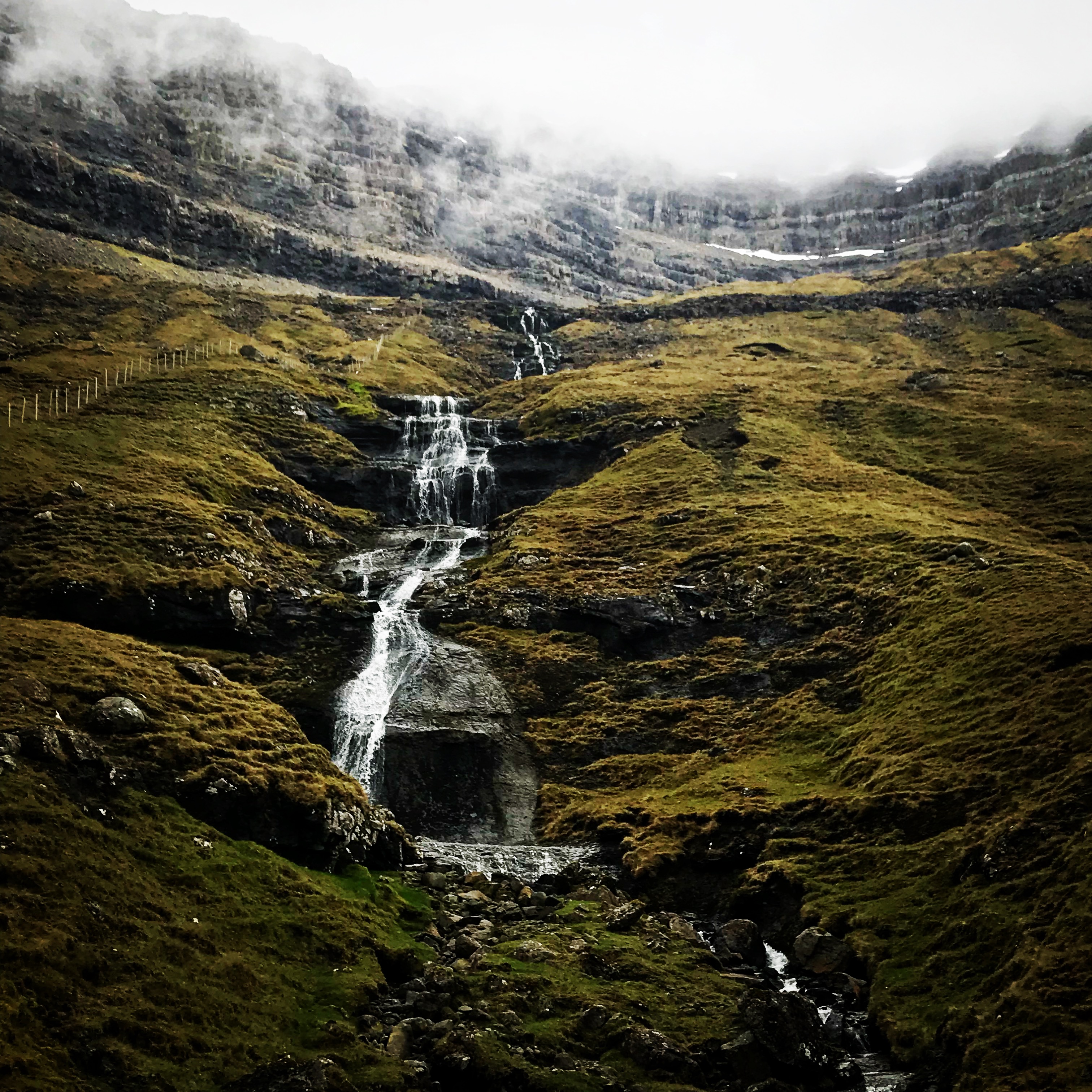 Bordoy, Faroe IslandsThis image showcases a cascading waterfall flowing down a mossy, rocky hillside in a remote and rugged landscape. The misty atmosphere and overcast sky add a dramatic and mysterious touch to the scene. The surrounding terrain, dotted with earthy greens and browns, emphasizes the natural beauty and isolation of this location.