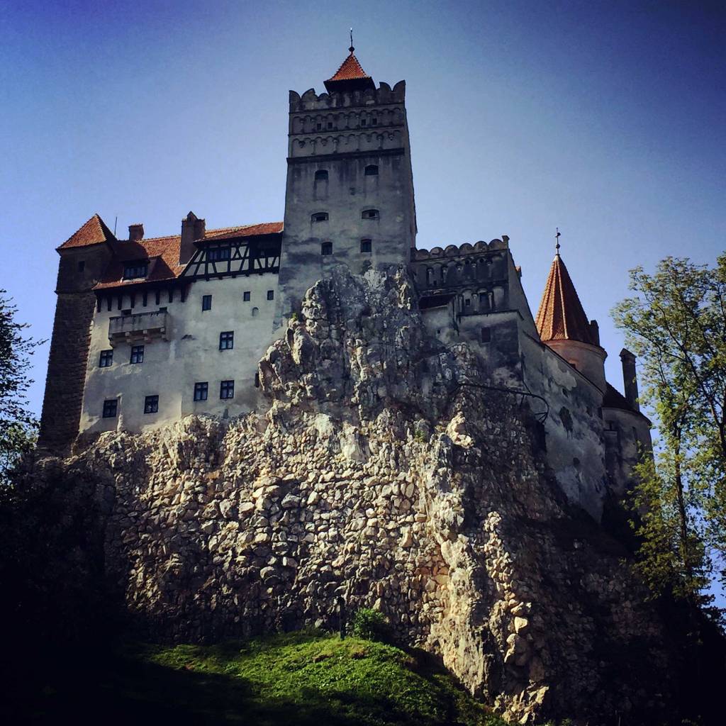 This image shows a dramatic view of a medieval castle atop a rocky cliff, with a mix of stone walls, pointed red rooftops, and arched windows. The scene is framed by trees and bathed in a contrast of shadow and sunlight under a clear blue sky.