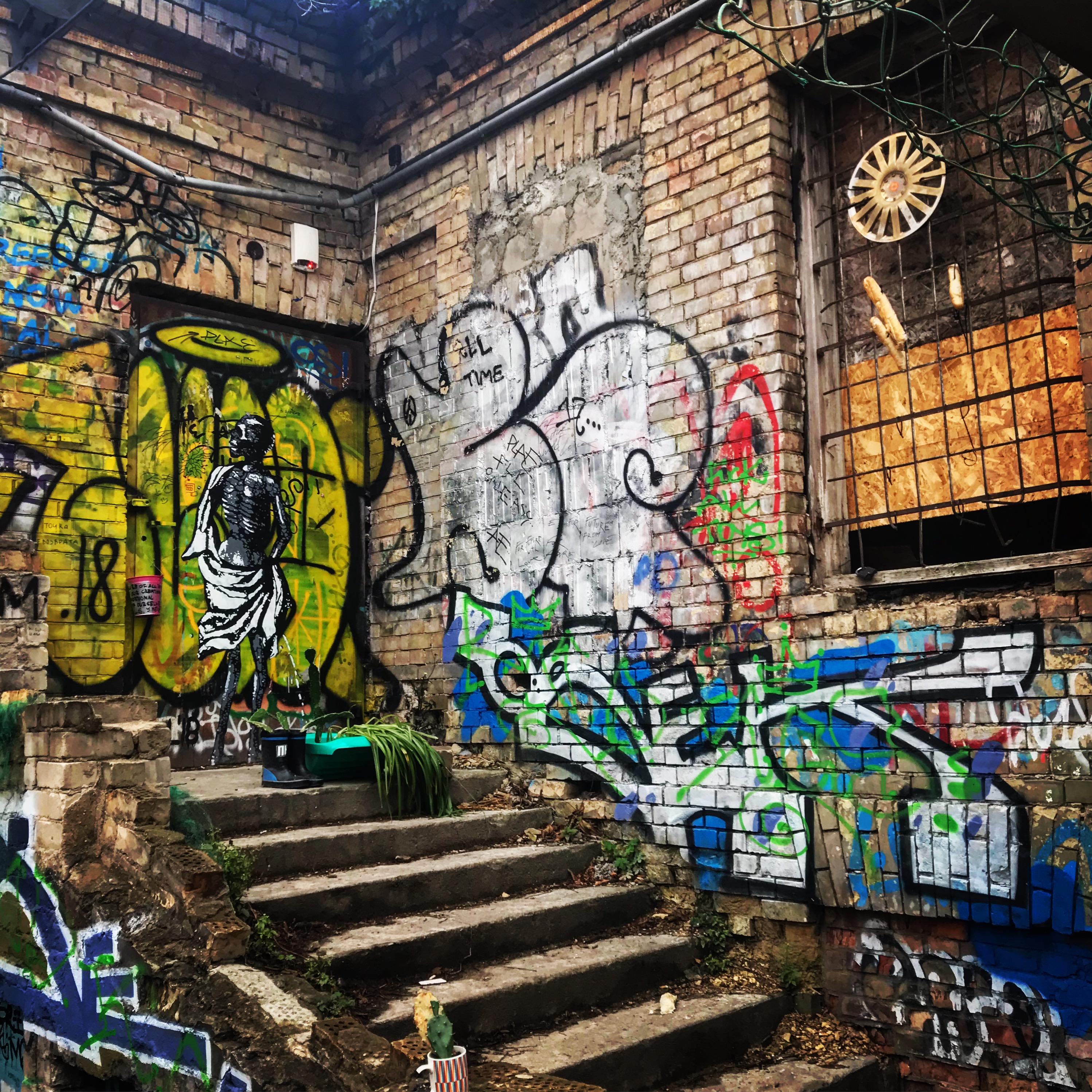 This image showcases a dilapidated brick corner adorned with eclectic graffiti, including a striking stencil of a skeletal figure draped in robes over a vivid yellow background. Crumbling stairs lead to the area, surrounded by vibrant tags and murals, while an old window with bars displays whimsical items like a wheel and dangling objects. The scene exudes a gritty yet artistic urban vibe.