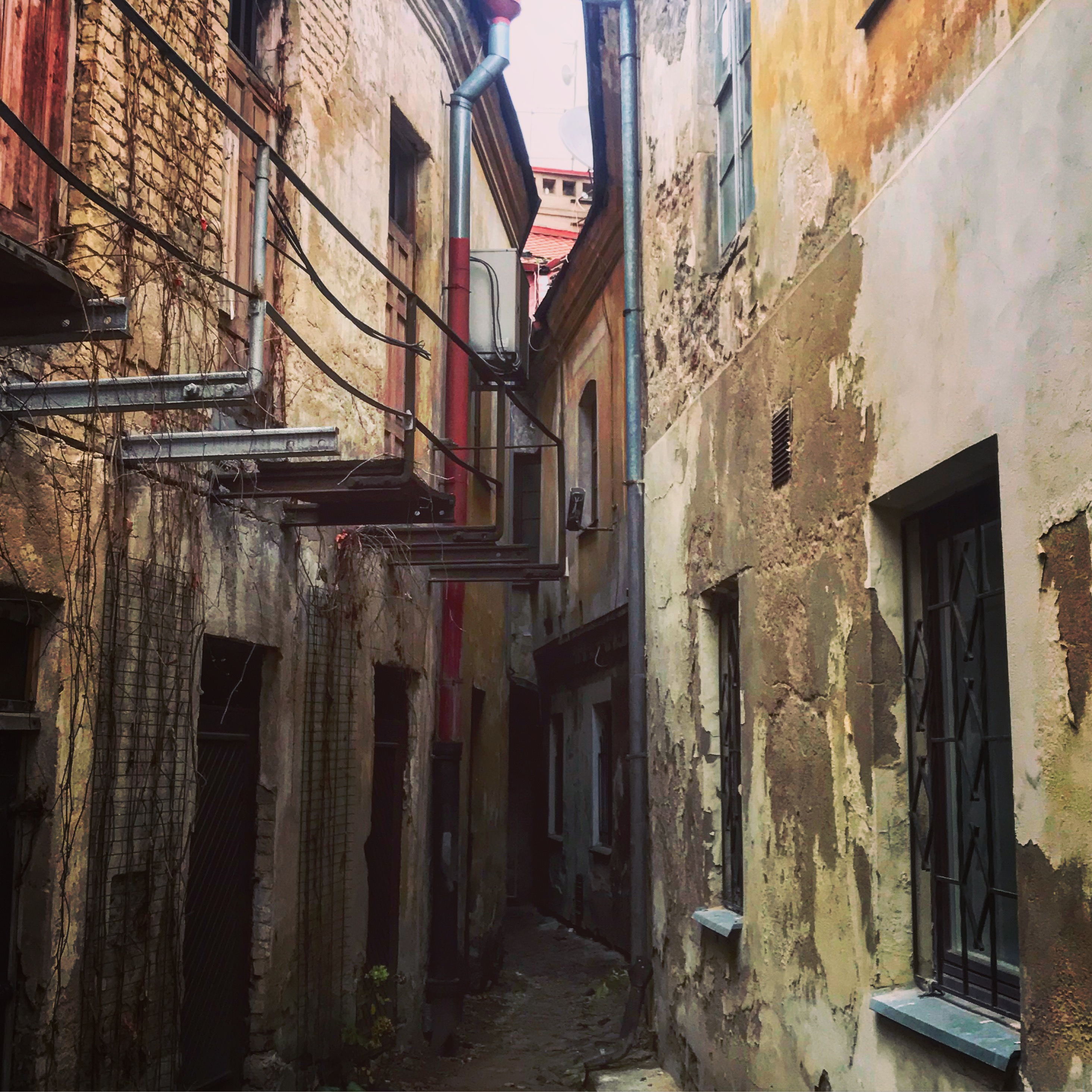 This image depicts a narrow alleyway between two aged, weathered buildings with peeling paint and exposed brickwork. Overhead, tangled wires and pipes add to the sense of urban decay, while faint light at the alley's end suggests depth and mystery. Let me know if you need further edits or specific details!