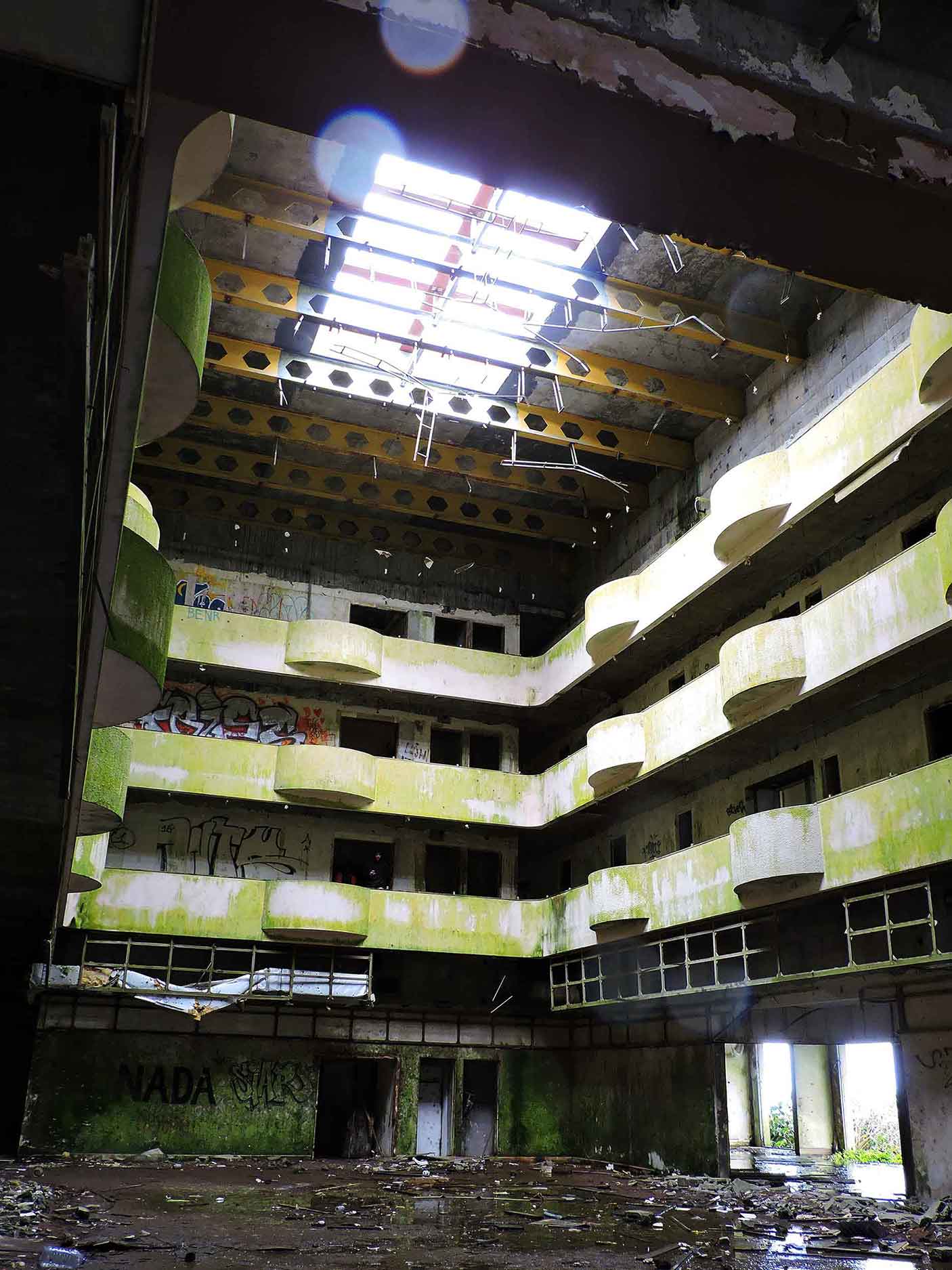 This image depicts the interior of an abandoned, multi-level building with moss and grime covering the curved balconies and walls, giving it a post-apocalyptic feel. Light streams through a broken skylight in the roof, highlighting scattered debris and graffiti on the dilapidated structure. The eerie mix of decay and partial illumination creates an atmosphere of haunting beauty.