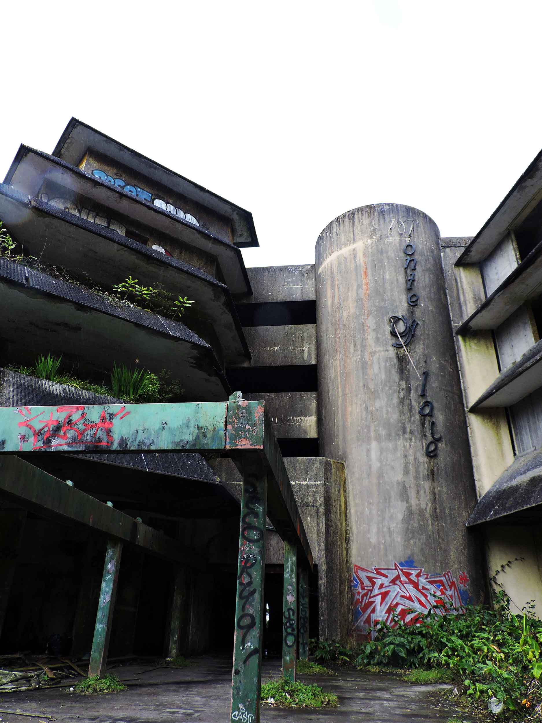 This image shows an abandoned, brutalist-style building with overgrown vegetation reclaiming its structure. The concrete walls are adorned with graffiti, adding an urban decay aesthetic, while the cylindrical tower prominently features faded text and marks of wear. The tilted beams and neglected state contribute to a haunting yet visually compelling atmosphere.