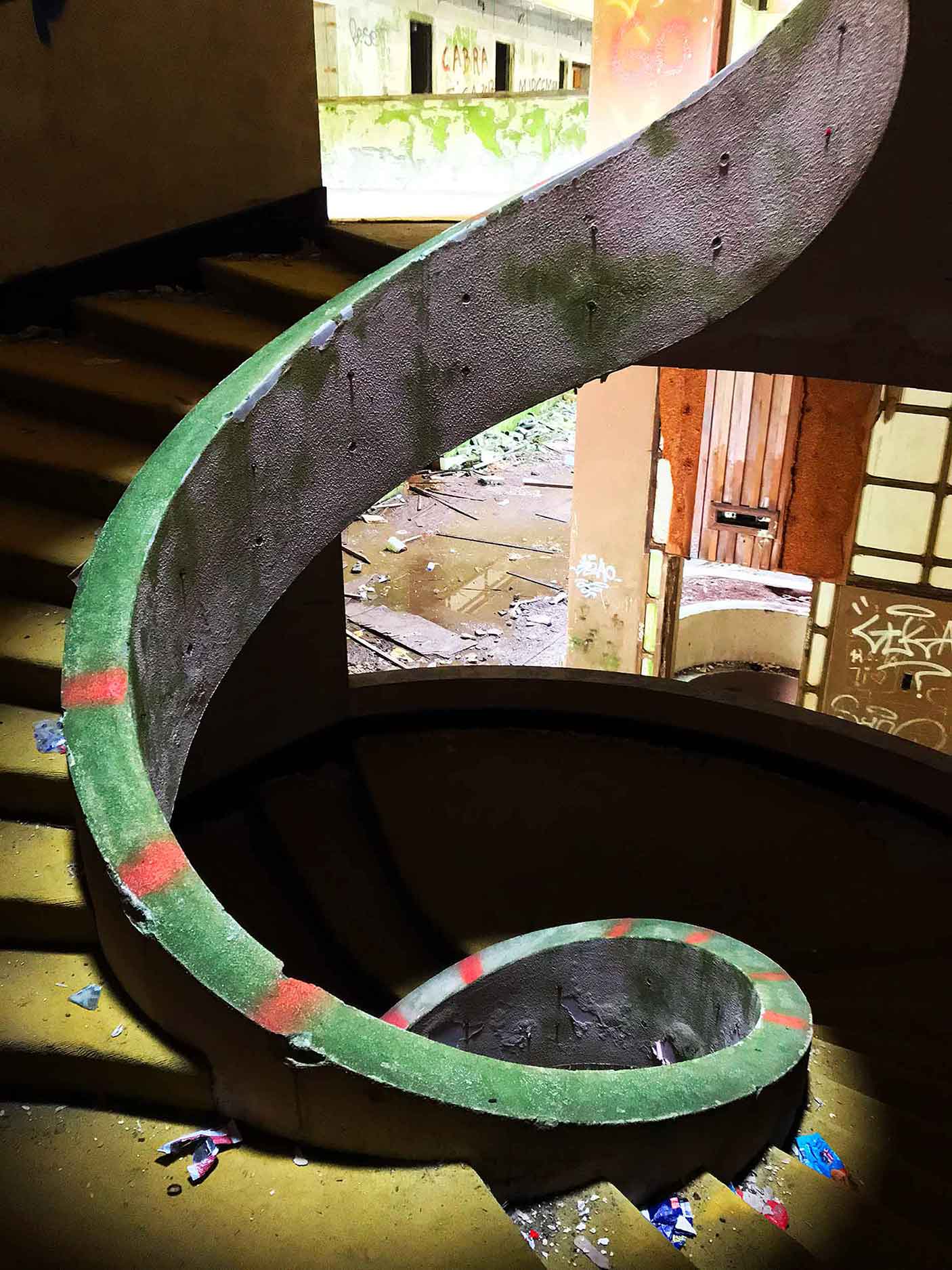 This image features a spiral staircase in an abandoned building, with a weathered green railing accented by faded red stripes. The space is dimly lit, revealing scattered debris and broken tiles on the stairs and floor below. The curved design contrasts with the decaying surroundings, creating an intriguing mix of elegance and neglect.