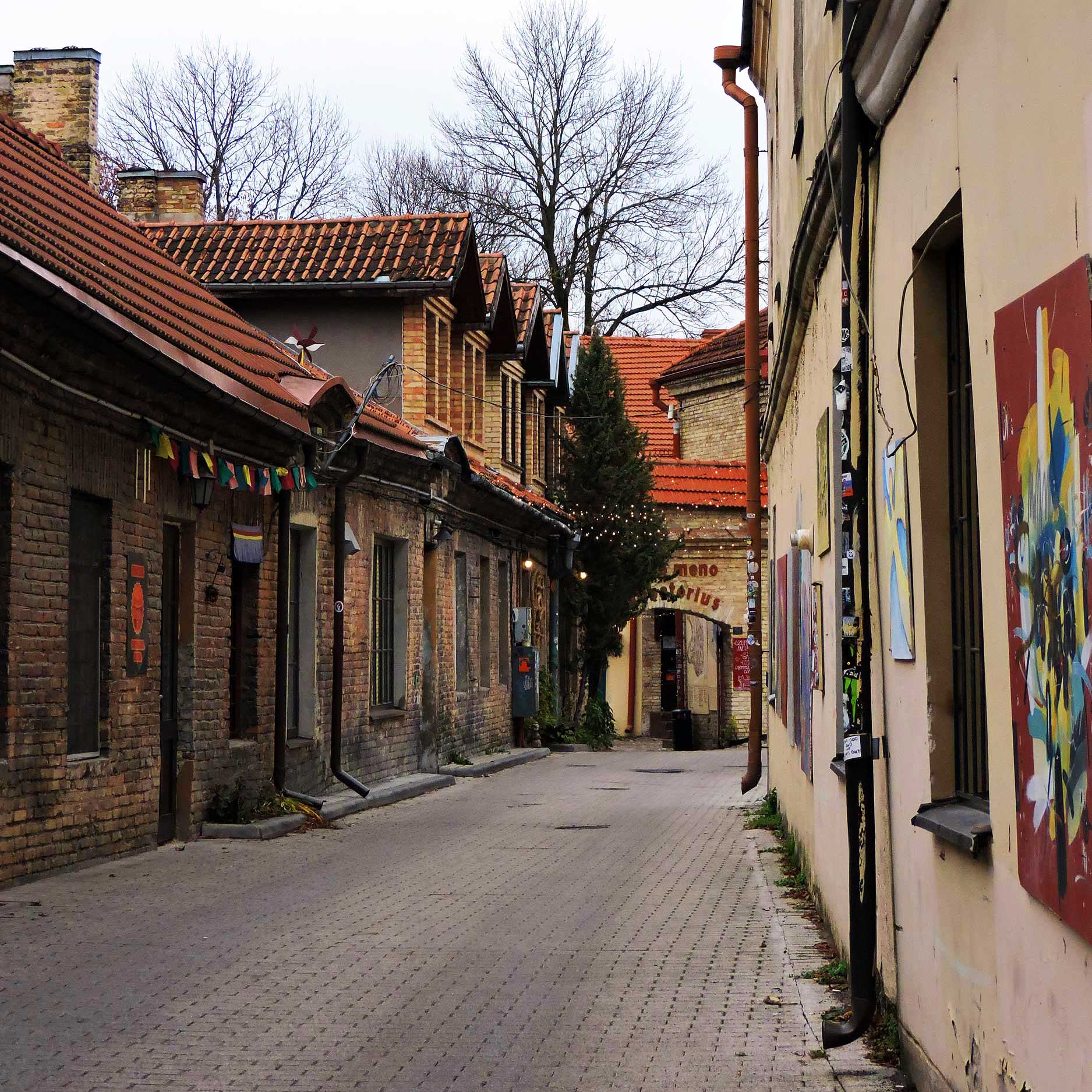 This image shows a charming cobblestone street flanked by old brick and plaster buildings with red-tiled roofs, adorned with colorful decorations and artwork. The scene leads to an archway in the distance, framed by bare winter trees and string lights, adding a cozy, artistic vibe. Let me know if youโd like any additional details!
