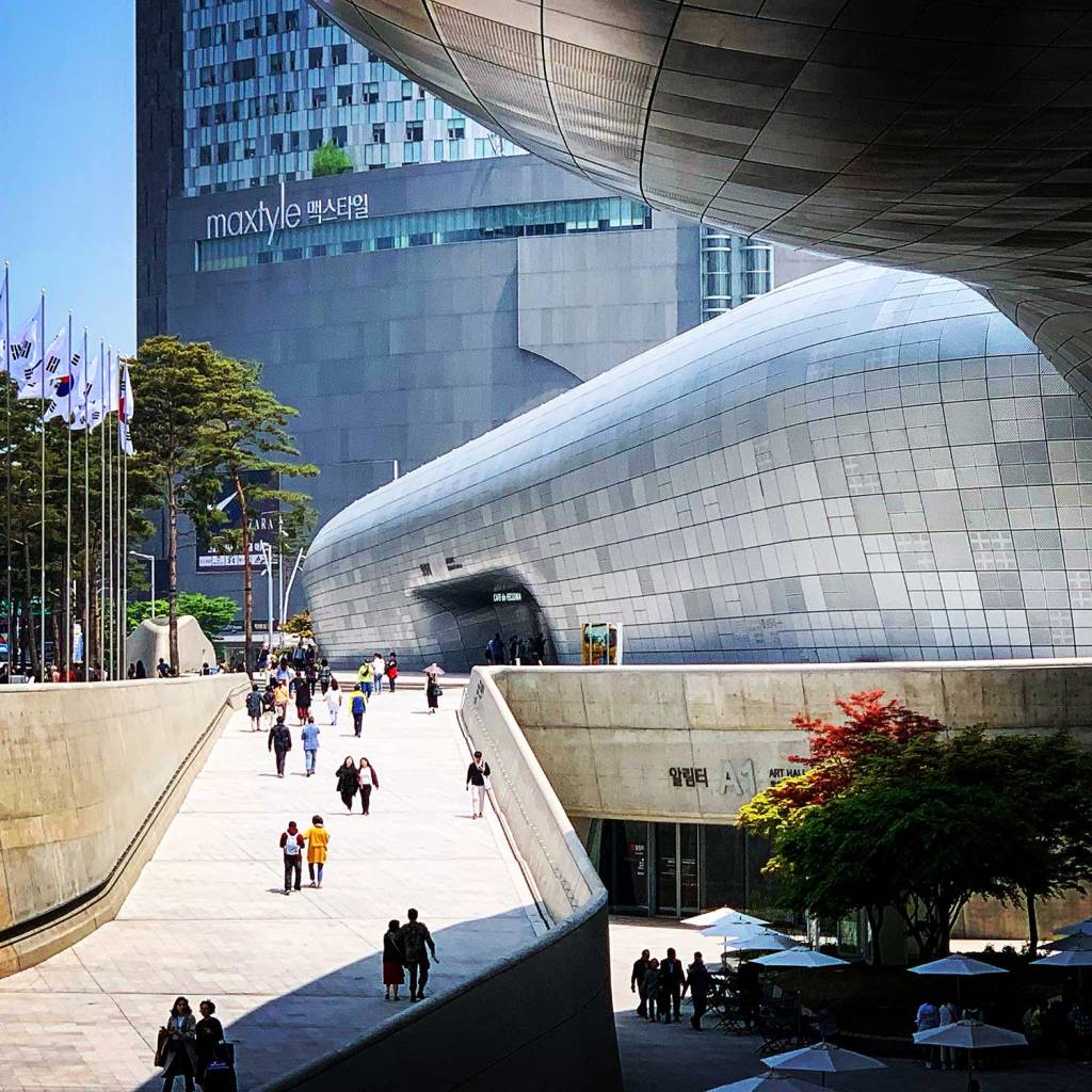 A futuristic urban scene at Dongdaemun Design Plaza in Seoul, with its sleek, curving silver architecture, a tree with red and green leaves in the courtyard, and people walking along wide, open walkways under a clear blue sky.