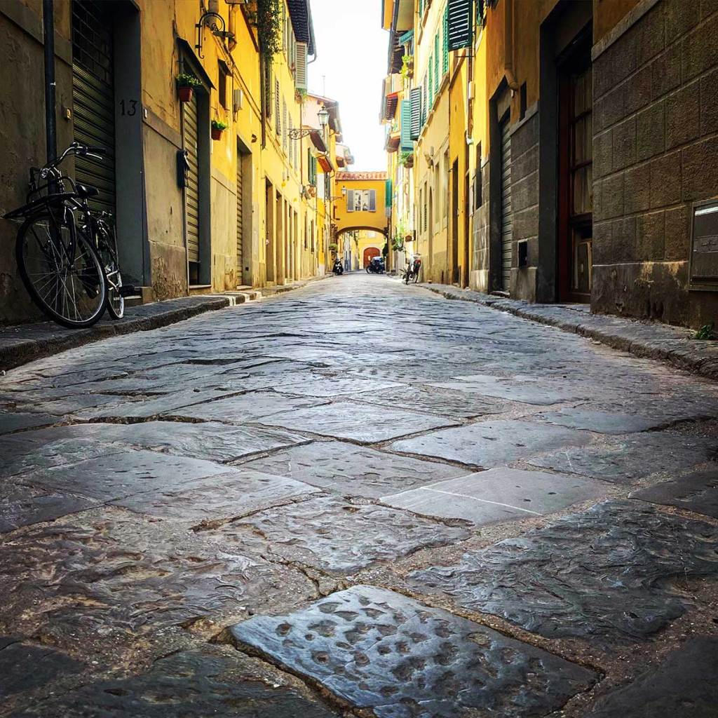 A cobblestone street in a historic Italian city, likely taken in Florence, lined with yellow and beige buildings. The image is taken from a low angle, emphasizing the texture of the cobblestones. The street is narrow and flanked by shuttered windows and doors, with a bicycle leaning against a wall on the left. In the distance, an archway frames the continuation of the street, leading further into the picturesque neighborhood. The overall mood is quiet and timeless, capturing the charm of a typical Italian alleyway.