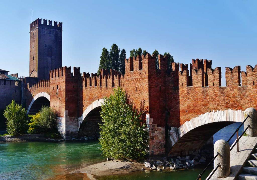 A view of the Castelvecchio Bridge, also known as the Scaliger Bridge, in Verona, Italy. The medieval bridge, constructed of red bricks, features crenellated battlements along its length, leading to the formidable Castelvecchio fortress in the background. The bridge arches gracefully over the Adige River, with green water flowing beneath it. A tall, square tower from the fortress rises prominently, adding to the imposing presence of this historic structure. The scene is bathed in bright sunlight, highlighting the rich red tones of the brickwork against a clear blue sky.