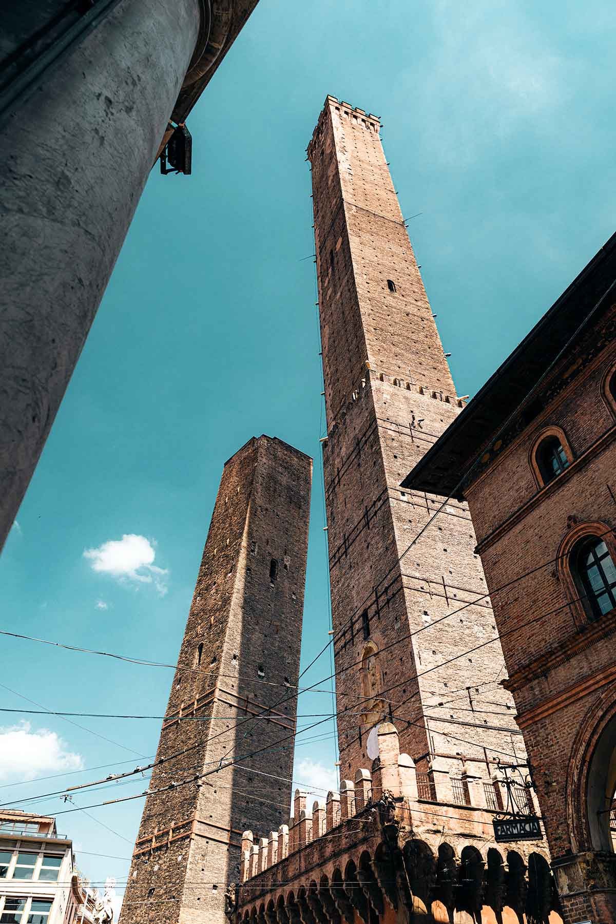 A dramatic upward view of the Two Towers, or "Le Due Torri," in Bologna, Italy. The image captures the taller Asinelli Tower and the shorter, leaning Garisenda Tower against a backdrop of a clear blue sky. The towers, built in the medieval period, are iconic symbols of Bologna. The surrounding buildings and electrical wires crisscrossing the scene add to the urban feel, while the historical architecture contrasts with the modern elements in the lower part of the image. The perspective emphasizes the height and grandeur of these ancient structures.