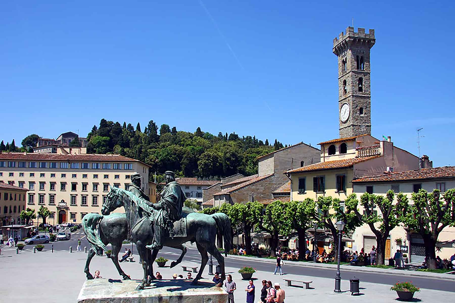 A vibrant scene from a town square in Fiesole, Italy, featuring a bronze equestrian statue in the foreground. The square is surrounded by historical buildings with red-tiled roofs, and the prominent clock tower rises against a backdrop of green hills and a clear blue sky. People are gathered around the square, enjoying the sunny day, with some sitting under the shade of neatly trimmed trees lining the street. The image captures the charm and historic atmosphere of this picturesque Italian town.