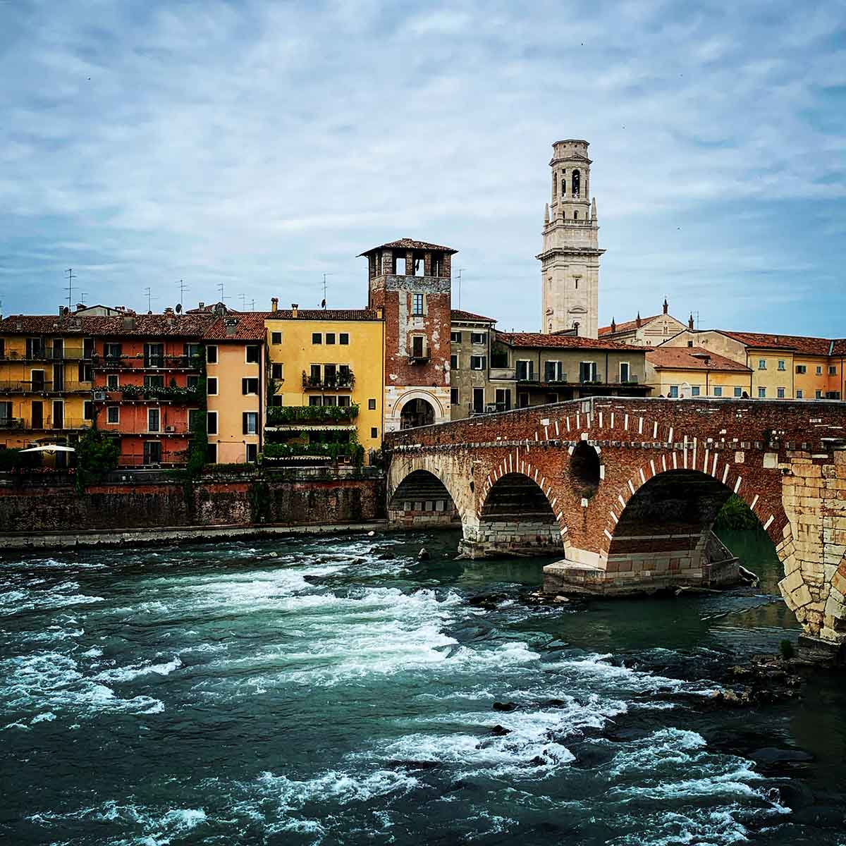 A scenic view of the historic Ponte Pietra in Verona, Italy, crossing the Adige River. The stone and brick bridge, with its arched design, stands prominently over the flowing waters below. On the opposite bank, colorful buildings with red-tiled roofs, including a yellow and orange faรงade, create a charming backdrop. A tall bell tower rises above the rooftops, adding to the picturesque skyline of Verona. The sky is overcast, giving the scene a moody yet timeless atmosphere, emphasizing the rich history and beauty of the location.