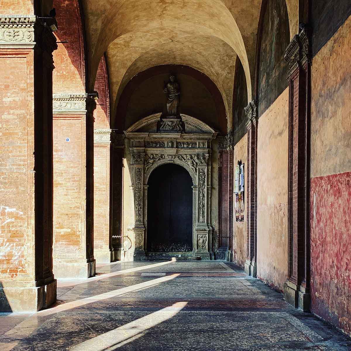 A serene and historic arcade in Bologna, Italy, with warm sunlight casting long shadows across the patterned stone floor. The walls are adorned with a mix of aged plaster and exposed brick, leading to a richly detailed archway at the end of the corridor. Above the arch, a statue stands in a recessed niche, adding to the sense of antiquity and reverence. The overall atmosphere is quiet and contemplative, capturing the timeless beauty of Bologna's architectural heritage.