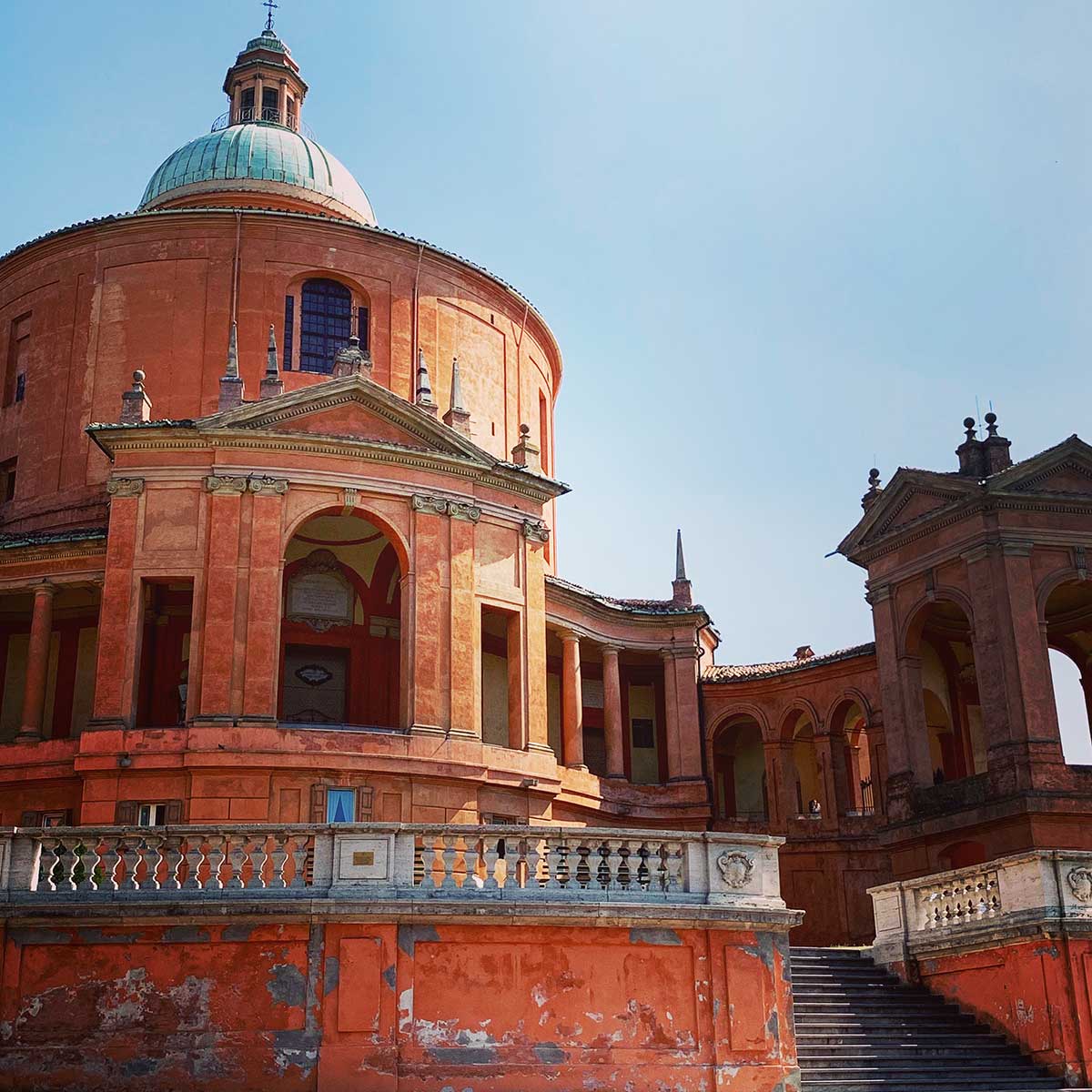 A view of the Sanctuary of the Madonna di San Luca in Bologna, Italy. The image captures the striking orange-pink faรงade of the church, with its large central dome and surrounding portico. The architectural details include arched entryways, stone balustrades, and a staircase leading up to the entrance. The buildingโs worn exterior adds to its historical charm, with the sunlight illuminating the vibrant colors against a clear blue sky. This sanctuary is a significant landmark, perched on a hill overlooking the city.