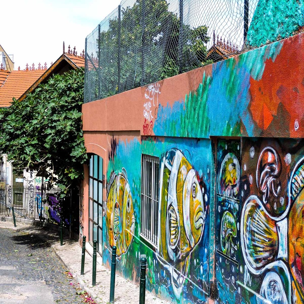 This image shows a vibrant mural painted on the side of a building in Lisbon, Portugal, featuring large, colorful fish in a stylized, underwater theme. The artwork covers a terracotta-colored wall, blending with the green and blue hues of the mural, creating a lively, artistic scene. The narrow, cobblestone street and lush greenery nearby add to the charming urban atmosphere of this creative corner of the city.