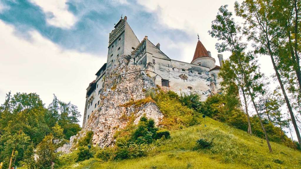This image depicts historic Bran Castle perched atop a rocky hill, surrounded by greenery and framed by tall trees under a partly cloudy sky. The architecture features a combination of towers and walls, blending into the rugged landscape.