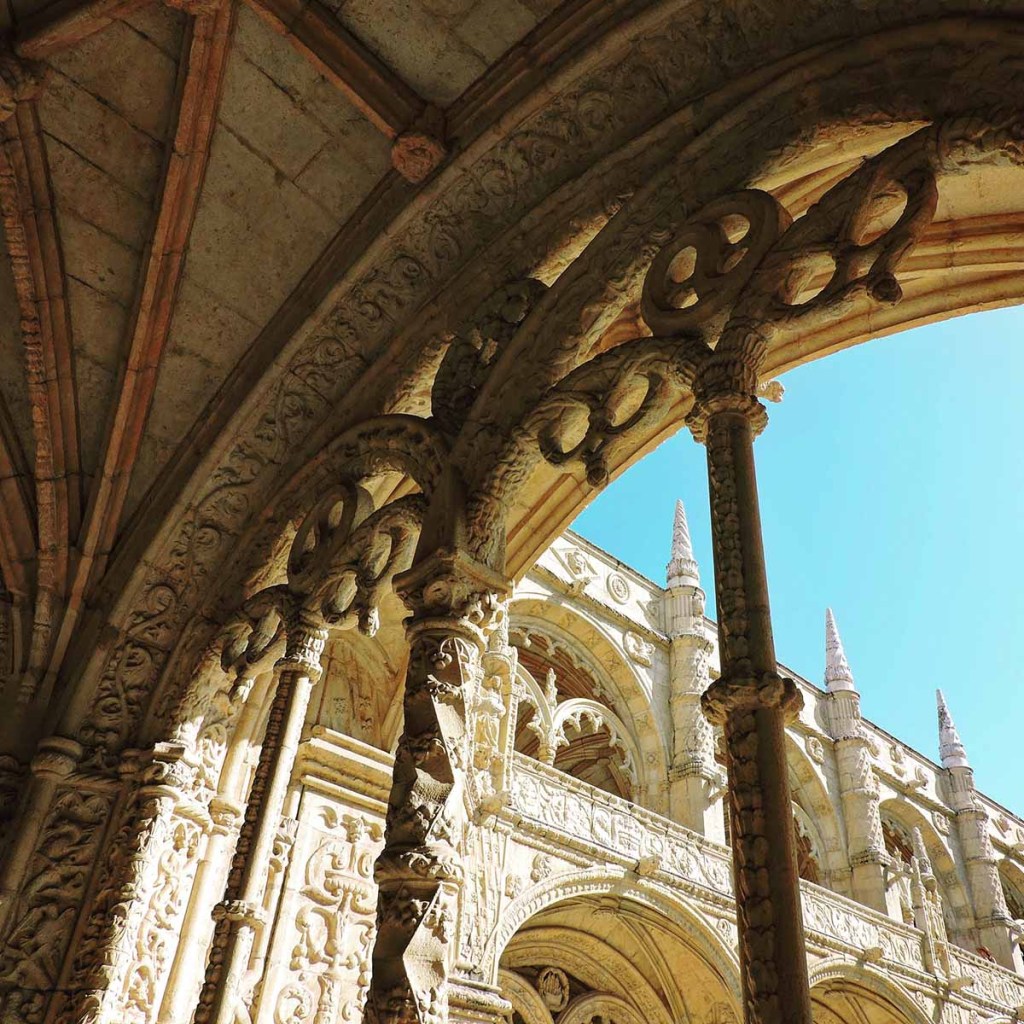 This image showcases the intricate stonework of the cloisters at Jerónimos Monastery in Lisbon, Portugal. The detailed arches and ornate carvings feature Gothic and Manueline architectural elements, highlighted by the warm sunlight against a clear blue sky. The elaborate design reflects the monastery’s historic grandeur and artistic craftsmanship.