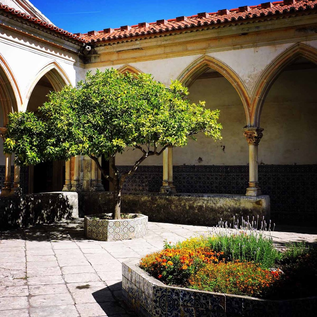 This image captures a serene courtyard within a monastery in Lisbon, Portugal, featuring Gothic arches and a central orange tree in a tiled planter. The lush greenery contrasts beautifully with the historic stone architecture and patterned azulejo tiles along the walls. Bright flowers add a touch of color, enhancing the peaceful, reflective atmosphere of this tranquil space.