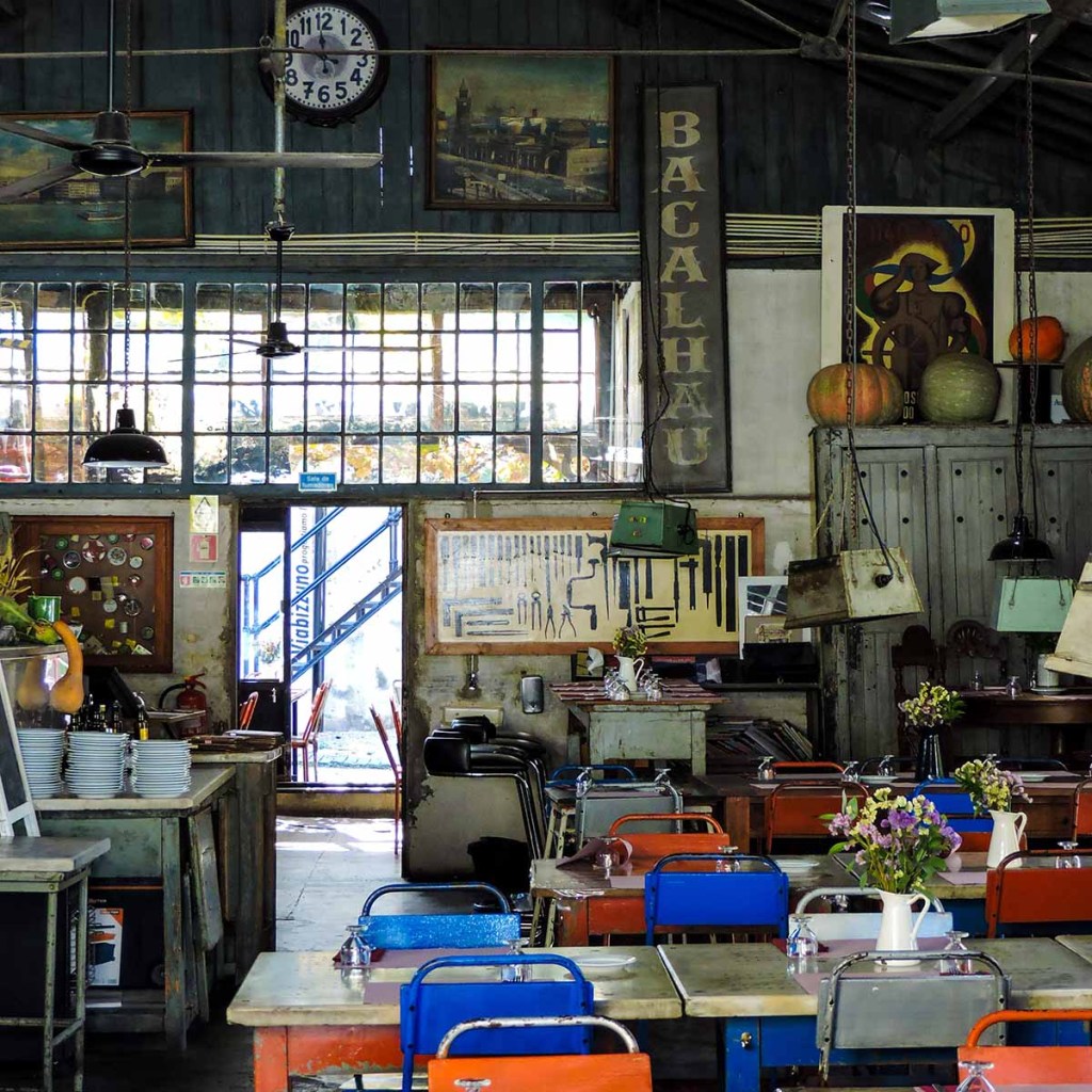 This image shows the interior of a quirky, vintage-style café or restaurant in Lisbon, Portugal. The space is filled with mismatched colorful chairs, retro decorations, and various antique items hanging on the walls, including a large sign reading “Bacalhau.” The eclectic decor, featuring old paintings, clocks, and industrial light fixtures, creates a nostalgic and unique ambiance reminiscent of a bygone era.