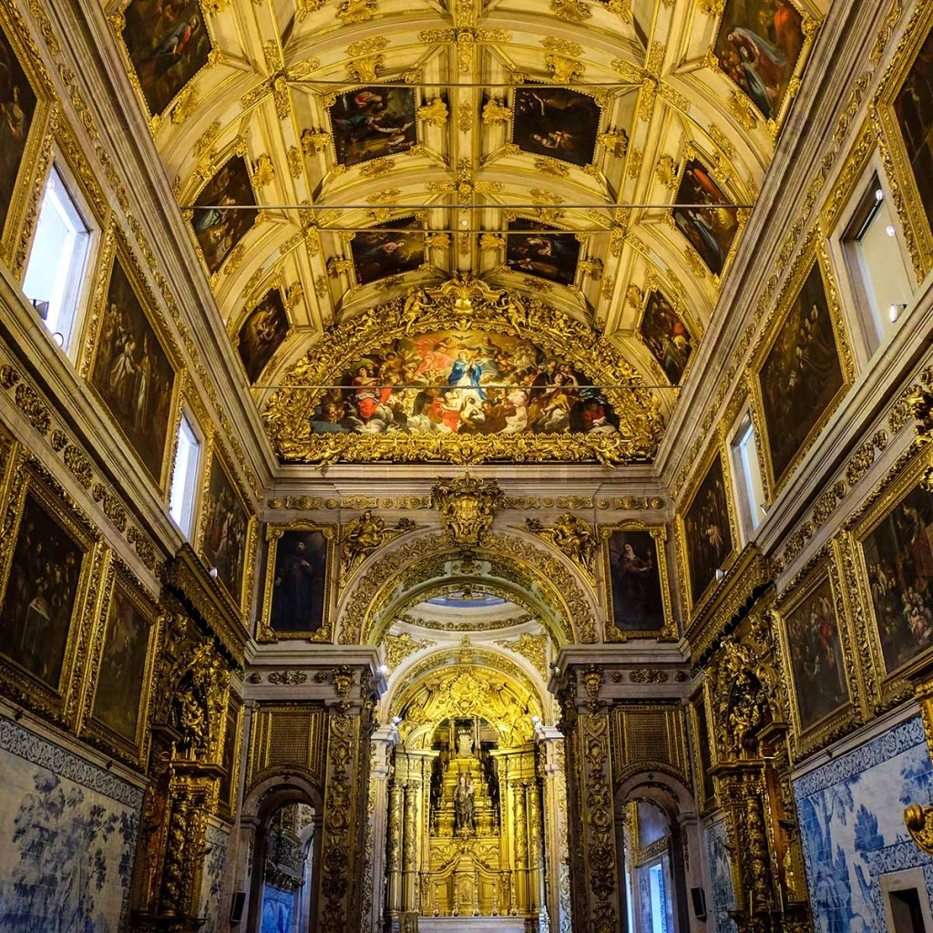 This image captures the opulent interior of a Baroque church in Lisbon, Portugal, adorned with gilded altars, ornate carvings, and richly painted ceilings. The walls are lined with framed religious artwork and detailed azulejo tiles, adding to the grandeur of the space. The ceiling's intricate paintings and gold accents reflect the church's historical and artistic significance, showcasing exquisite craftsmanship and religious iconography.