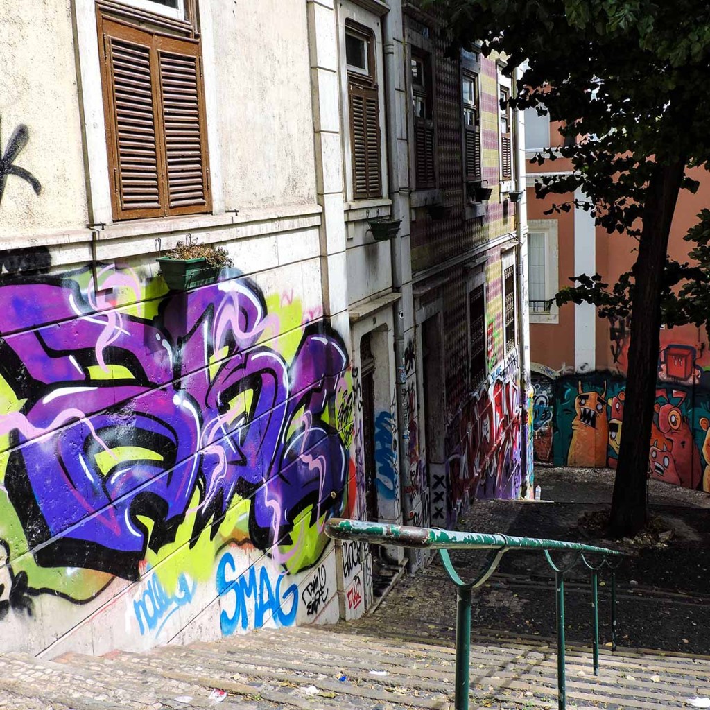 This image shows a narrow, graffiti-covered staircase in Lisbon, Portugal, descending between old buildings with shuttered windows. The walls are filled with colorful street art, including bold purple and green tags that add an edgy, urban feel to the scene. The steps are shaded by trees, creating a gritty yet artistic alleyway characteristic of Lisbon's vibrant street culture.