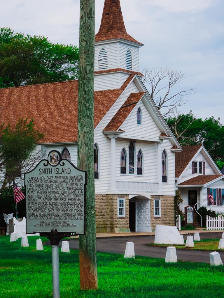 A historical marker for Smith Island stands in front of a charming white church with a brown shingle roof. The sign provides information about the island's history and early settlers, while the church's architecture, including its steeple and stained glass windows, adds to the quaint and historical ambiance of the scene.