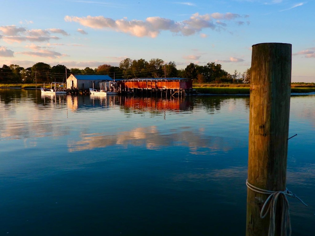 A tranquil waterside scene featuring a small dock with boats tied up next to rustic buildings, reflecting in the calm water under a sky with scattered clouds. A wooden post with a rope in the foreground adds depth to the picturesque setting.