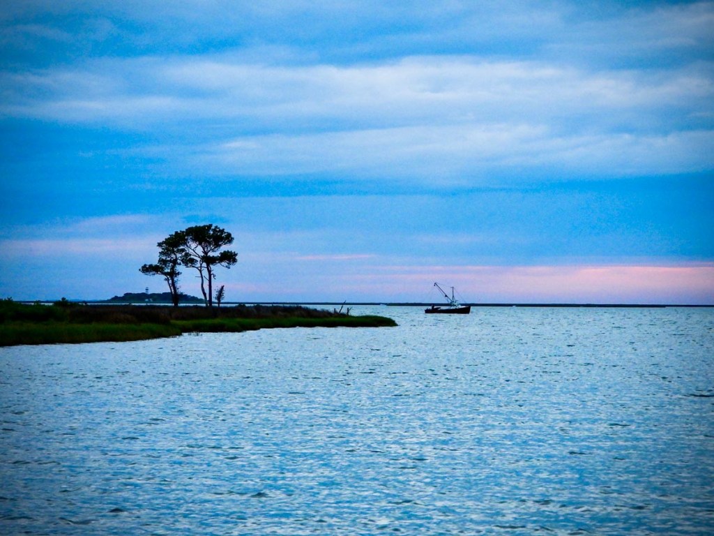 A serene coastal scene at dusk, featuring a solitary boat in the distance against a backdrop of a blue sky with hints of pink. A small patch of land with a few trees juts out into the water, adding a tranquil element to the expansive view of the sea.