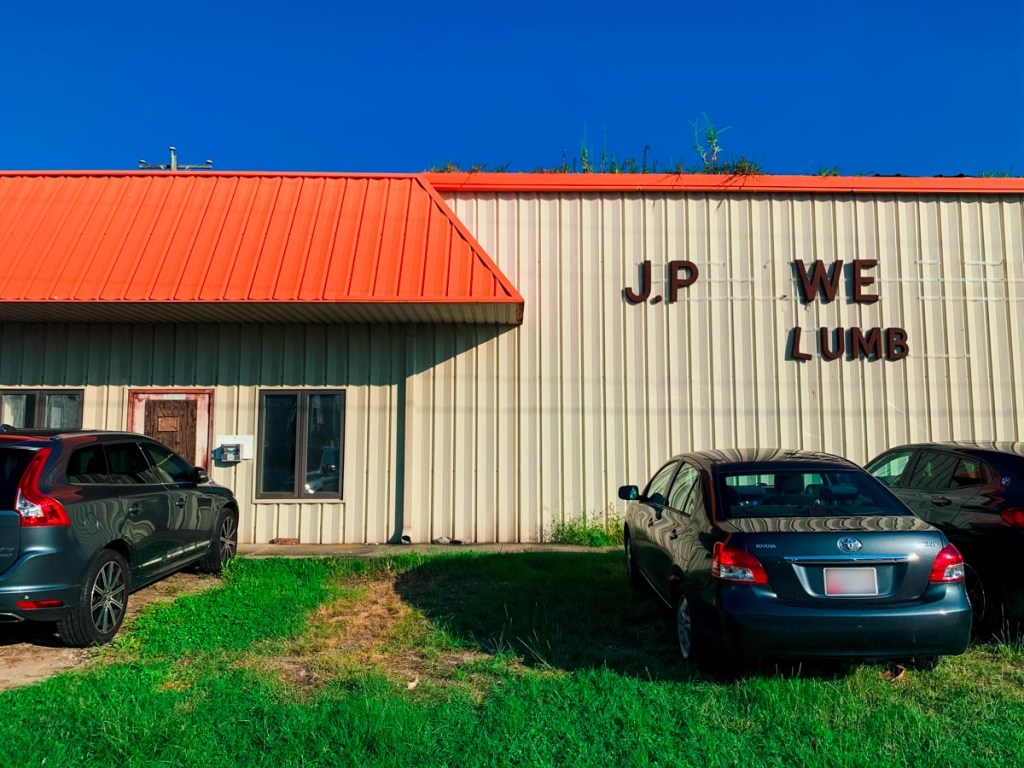 The exterior of a metal building with a red roof, partially showing the sign "J.P. WE LUMB." Two cars are parked in front on a patch of grass under a clear blue sky. The building appears to be a commercial property, possibly a lumber or hardware store.