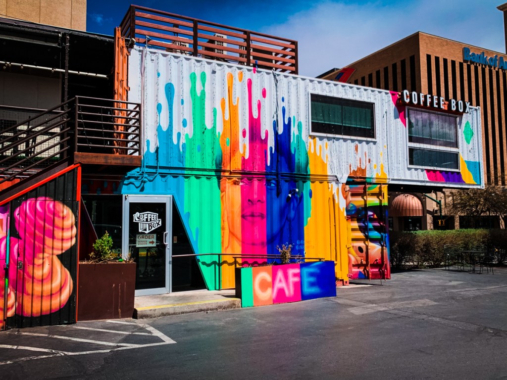 A vibrant café named "Coffee Box" is housed in a repurposed shipping container covered with colorful murals. Bold, dripping paint designs in shades of blue, green, yellow, pink, and orange decorate the exterior, creating a striking and artistic look. The word "CAFE" is painted in large, bright letters near the entrance. An outdoor staircase leads to an upper level, and a nearby building, marked with the "Bank of America" logo, is visible in the background. The scene captures a trendy, urban vibe.