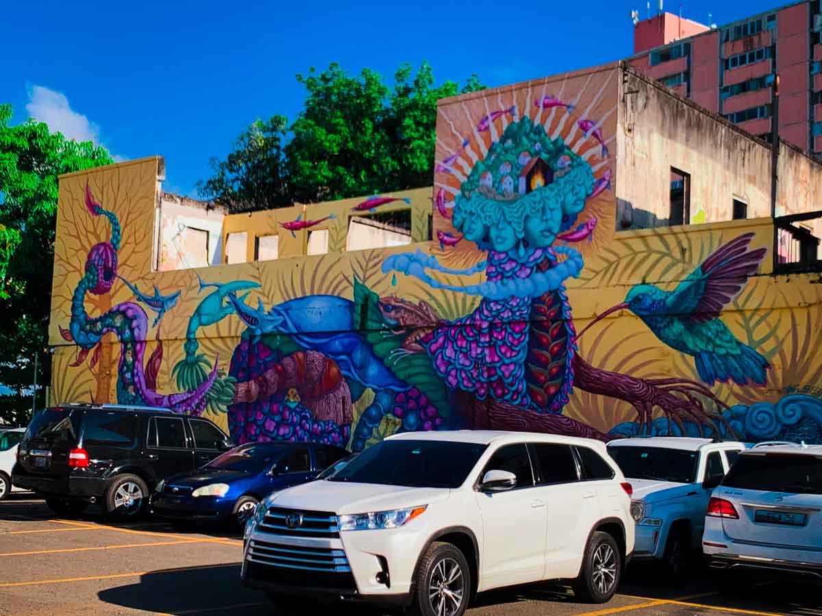 This image showcases a vibrant mural on the side of a building, featuring a fantastical scene with a mix of mythical creatures and nature, depicting a rally cry to save the planet. The mural includes a large central figure resembling a multi-headed creature adorned with floral patterns and surrounded by swirling, colorful elements such as snakes, a large bird, and other abstract shapes. The artwork is set against a yellow background, which contrasts with the vivid blues, purples, and greens of the figures. Below the mural, several cars are parked in a lot, giving a sense of scale to the expansive and detailed artwork. The scene is framed by a bright blue sky and lush green trees in the background.