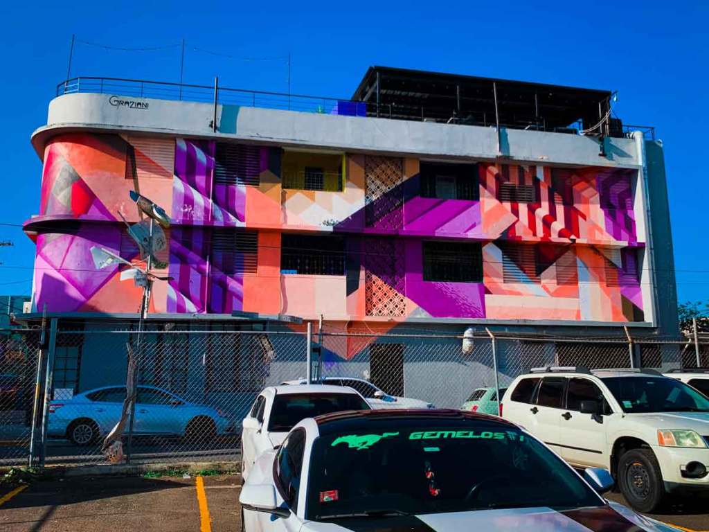 This image captures a building adorned with a vibrant geometric mural, featuring a mix of pink, purple, orange, and red hues. The design consists of overlapping shapes and patterns that create a sense of depth and movement across the facade. The building's curved corners and flat surfaces are seamlessly integrated into the mural, enhancing its three-dimensional effect. Below the mural, several cars are parked in a lot, and the area is enclosed by a chain-link fence. The name "Graziani" is visible on the top left corner of the building, likely indicating the artist or the building’s name. The bright blue sky provides a striking contrast to the bold colors of the mural, making the artwork stand out in the urban landscape.