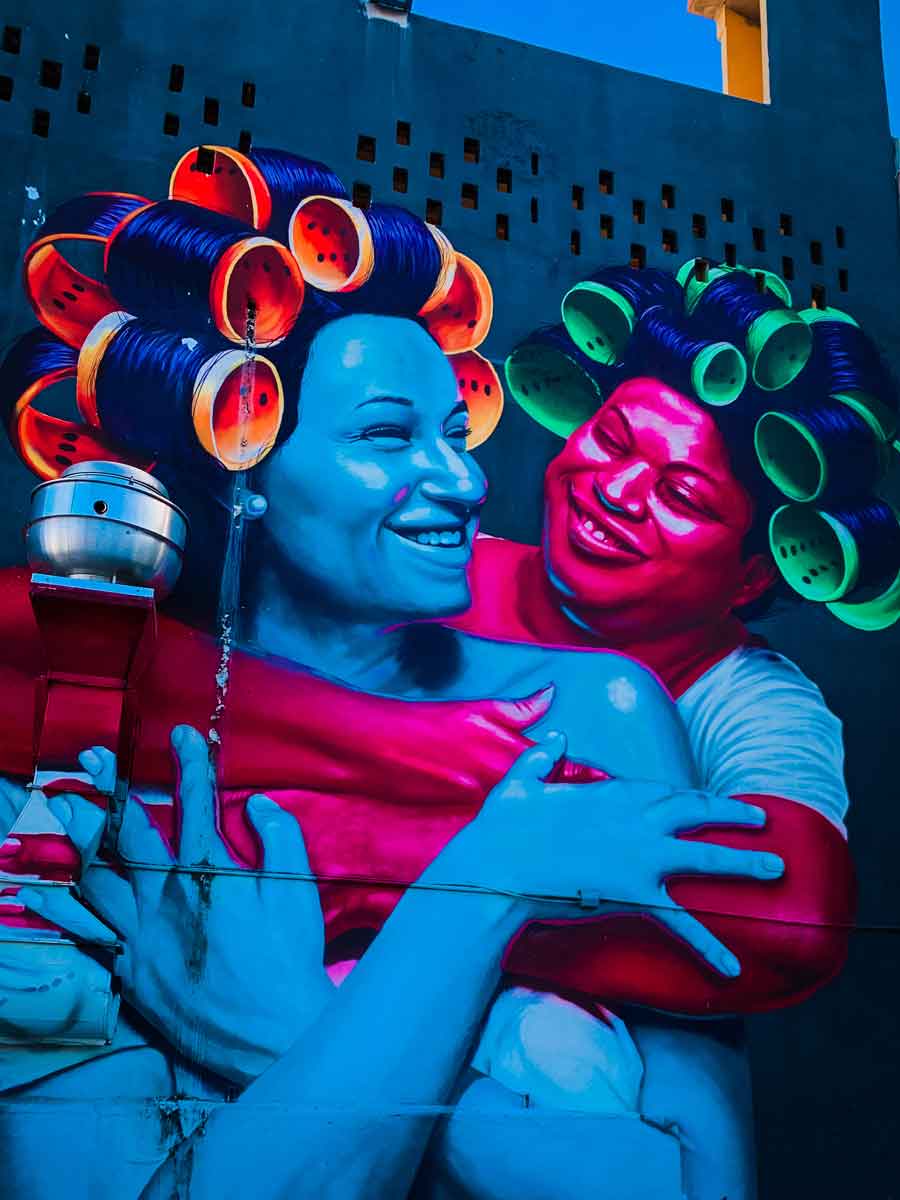 This image displays a vibrant mural of two women embracing, both smiling warmly at each other. The figures are painted in bold, contrasting colors, with one woman in shades of blue and the other in pink. They are depicted wearing large, colorful hair rollers, adding a playful and affectionate tone to the artwork. The background is a dark teal, which makes the bright colors of the figures pop even more. The mural conveys a sense of joy, intimacy, and connection between the two women, captured in a moment of shared happiness. The vivid colors and dynamic composition make the artwork stand out dramatically against the urban setting.