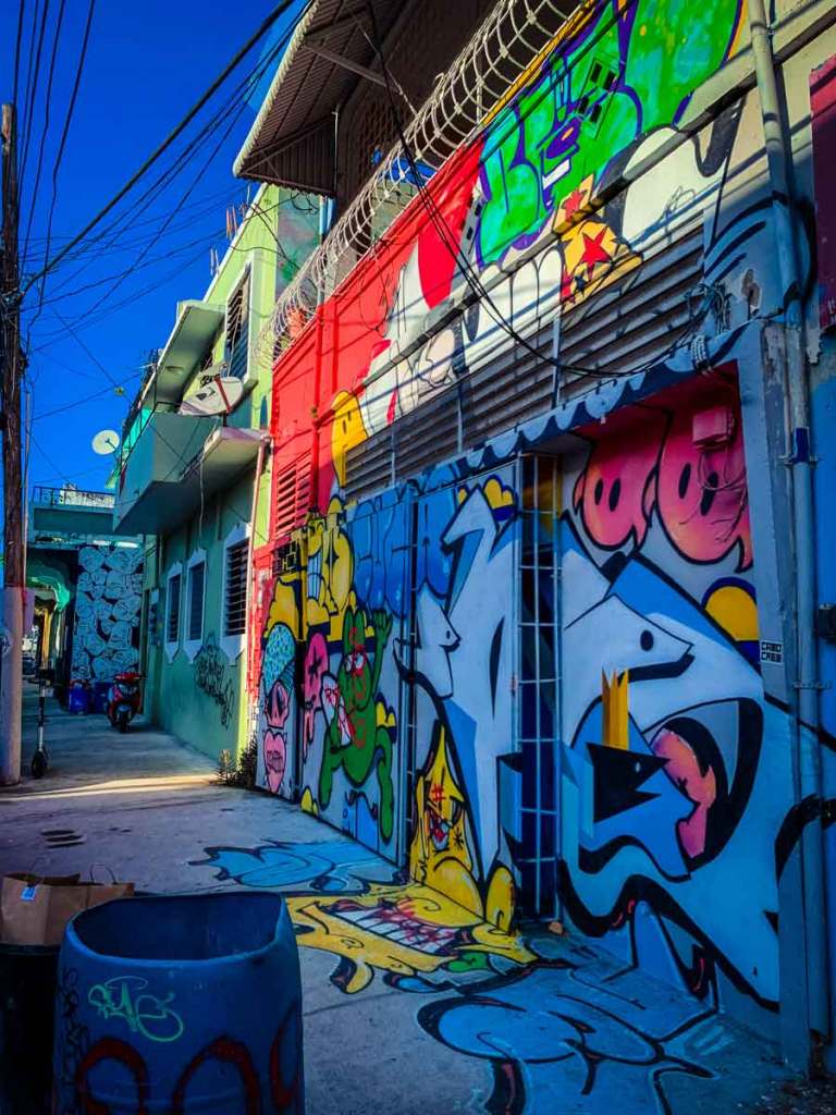 This image shows a narrow alleyway with walls covered in vibrant, colorful graffiti art. The artwork features various abstract designs and characters, blending multiple colors like blue, red, and green. Overhead, there are wires and barbed wire adding an urban, gritty feel to the scene, contrasting with the lively street art below.