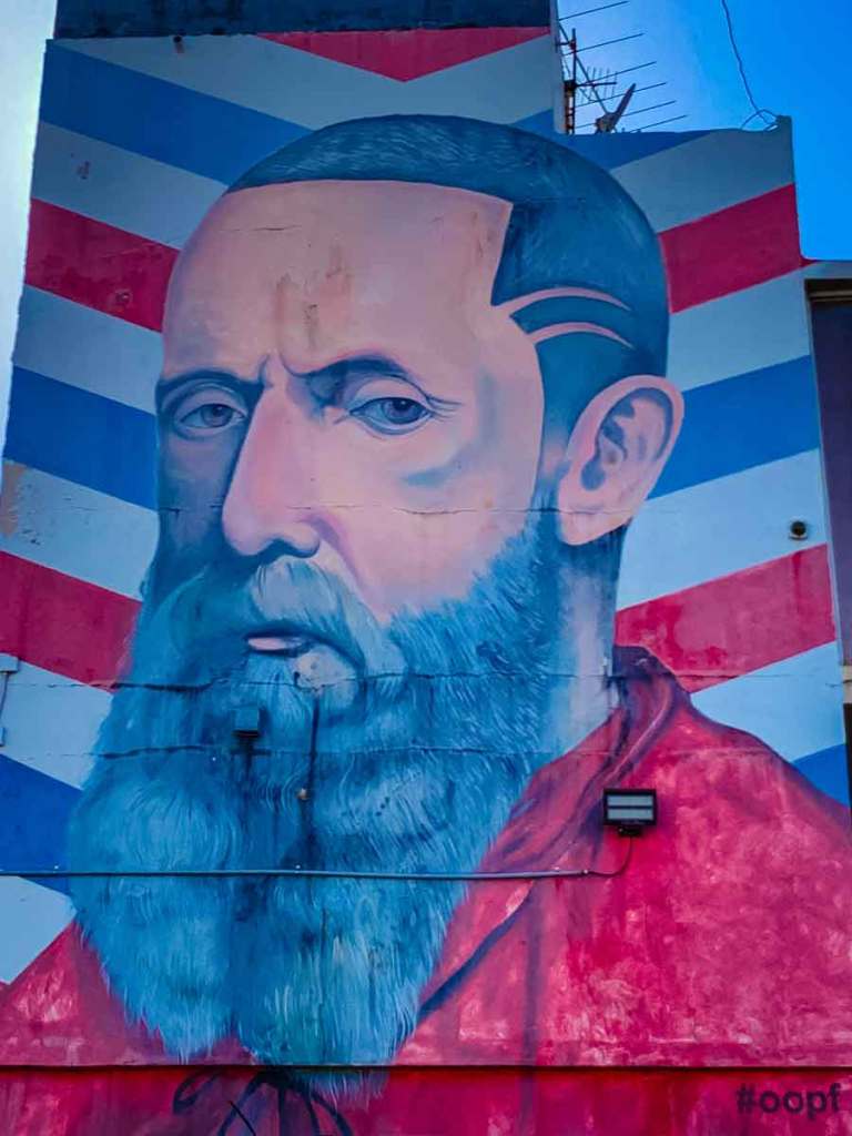 This image depicts a vibrant mural  in santurce san juan puerto rico, of a man with a blue beard and red robe, set against a background of diagonal red, white, and blue stripes. The man's expression is solemn, with detailed facial features, and there is a small hashtag "#oopf" in the lower right corner of the mural. The artwork appears to be painted on the side of a building.