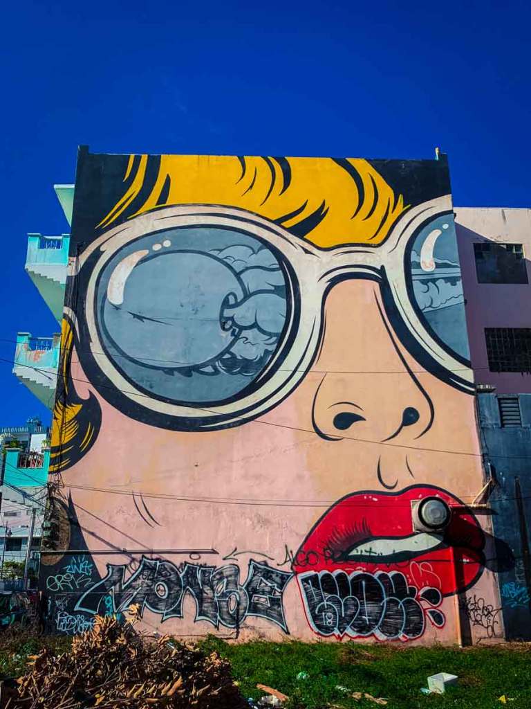 This image features a large, striking mural painted on the side of a building. The artwork depicts a close-up of a face, focusing on oversized sunglasses reflecting a cloudy sky and waves, with bright yellow hair and bold red lips. The lower part of the mural is tagged with graffiti, adding an additional layer of urban texture to the scene. The vibrant colors contrast sharply against the clear blue sky.