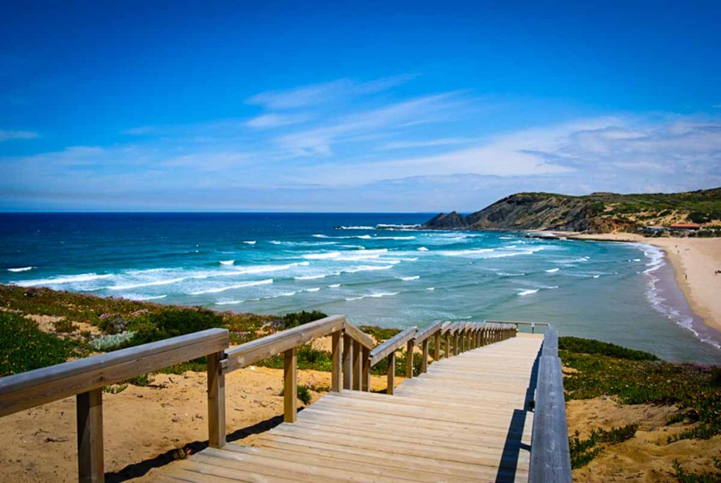 This image shows a beautiful coastal landscape in Portugal, featuring a wooden boardwalk leading down to a sandy beach with gentle waves rolling in. The turquoise waters of the ocean contrast with the golden sand and the green, rocky hills in the background, creating a serene and picturesque seaside view. The clear blue sky enhances the idyllic and inviting atmosphere of this scenic beach.