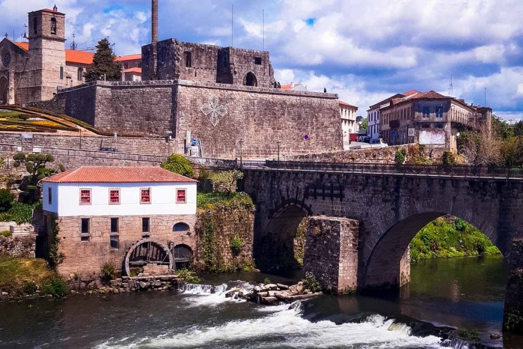 This image features a historic riverside scene in Portugal, with an ancient stone bridge arching over a flowing river. A medieval fortress with thick stone walls dominates the background, while a charming white building with a red-tiled roof sits by the water's edge, adding to the rustic charm. The setting is framed by lush greenery and traditional architecture under a partly cloudy sky, reflecting the rich history and scenic beauty of the area.