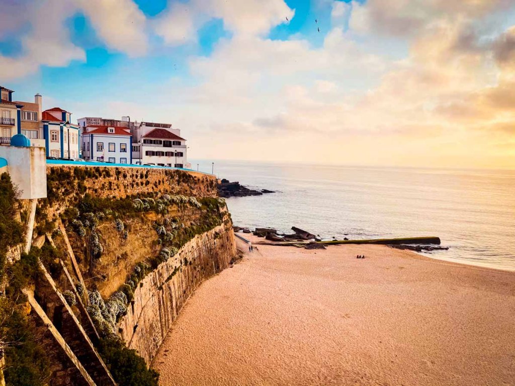 This image beautifully showcases a coastal town perched on cliffs overlooking a serene beach and ocean. The scene is bathed in warm, soft light, likely during sunrise or sunset, adding a golden hue to the sand and the buildings. The rugged cliffs, with greenery and old structures, give the area a quaint and picturesque feel, perfect for illustrating the allure of seaside travel destinations. This setting would be great for promoting slow travel or highlighting hidden coastal gems in Europe. If you need help integrating this image into your content, let me know how I can assist!