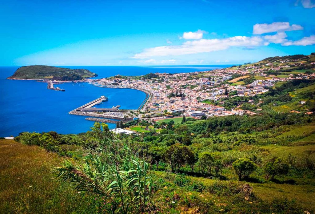This image captures a stunning coastal town nestled between the vibrant blue ocean and lush green hills. The view, taken from an elevated position, showcases a sprawling harbor with breakwaters and a mix of traditional and modern buildings spread along the coastline. The clear, sunny day highlights the contrast between the deep blue sea and the vivid green landscape, making it an ideal depiction of a scenic and tranquil coastal destination. This would be perfect for illustrating content about scenic overlooks, beautiful coastal towns, or the allure of slow travel to lesser-known seaside locations. Let me know if you need any assistance with using this image in your work!