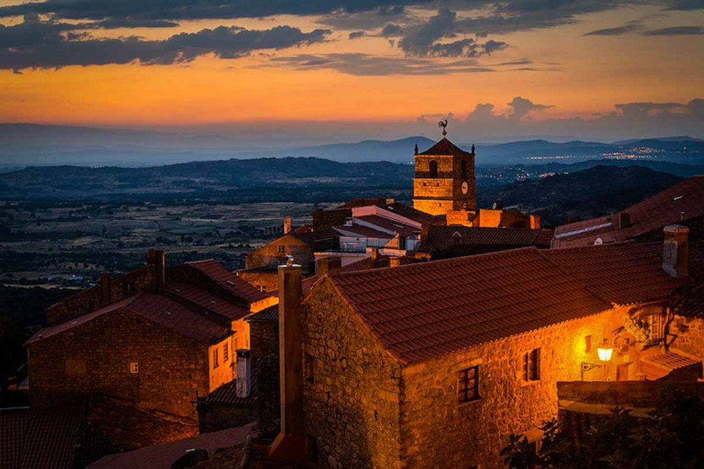 This image shows a stunning sunset over a historic town with glowing rooftops and a clock tower, set against a vast landscape. The warm, golden lighting creates a peaceful and timeless atmosphere.
