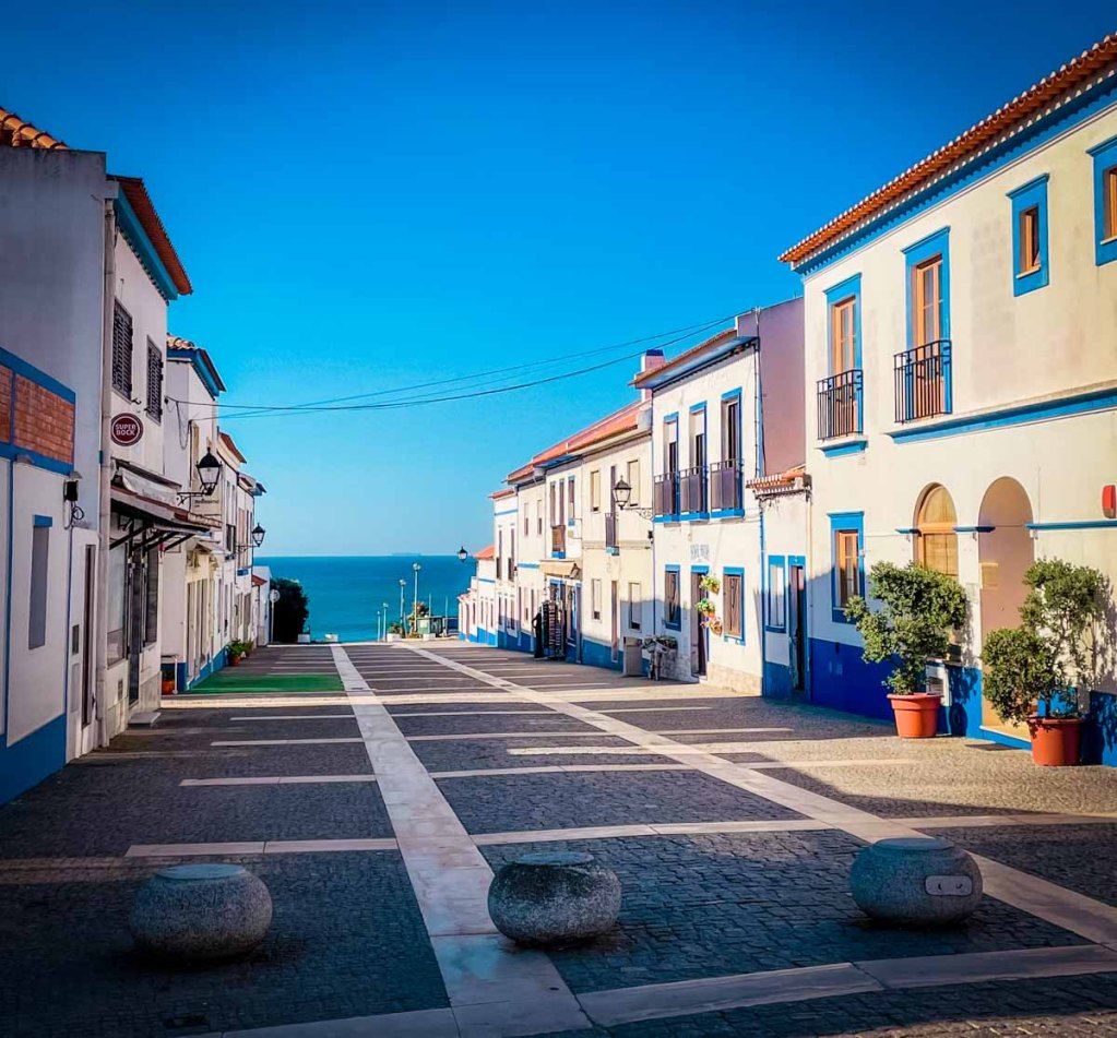 This image features a quiet, sunlit street lined with charming white and blue buildings leading to the sea. The clear blue sky and cobblestone path create a picturesque, serene setting typical of coastal European towns.