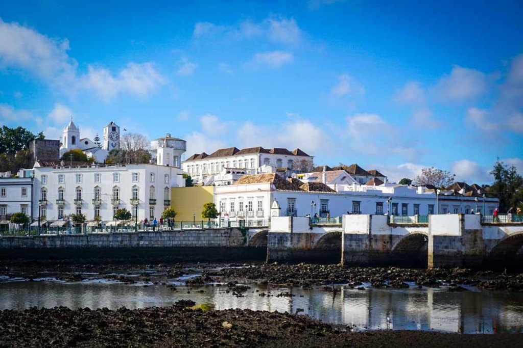 This image features a picturesque waterfront town with elegant white buildings, a historic clock tower, and a charming arched bridge. The bright blue sky adds to the serene and inviting ambiance of this scenic spot.