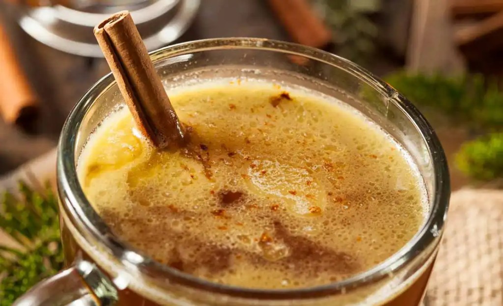 Close-up of a warm buttered rum cocktail garnished with a cinnamon stick, a cozy Friendsgiving drink idea. The frothy and spiced drink is served in a glass mug, creating a comforting and rich beverage perfect for a festive Friendsgiving gathering.