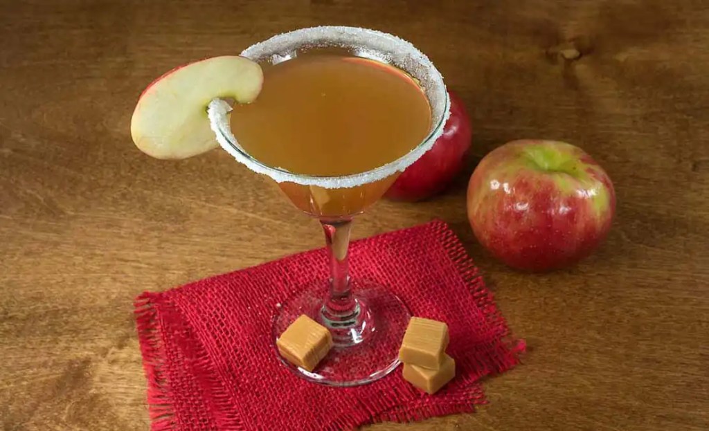 A caramel apple martini garnished with a fresh apple slice, an indulgent Friendsgiving drink idea. The cocktail is served in a sugar-rimmed martini glass on a red napkin, with apples and caramel pieces in the background, adding a sweet and festive touch to a Friendsgiving celebration.