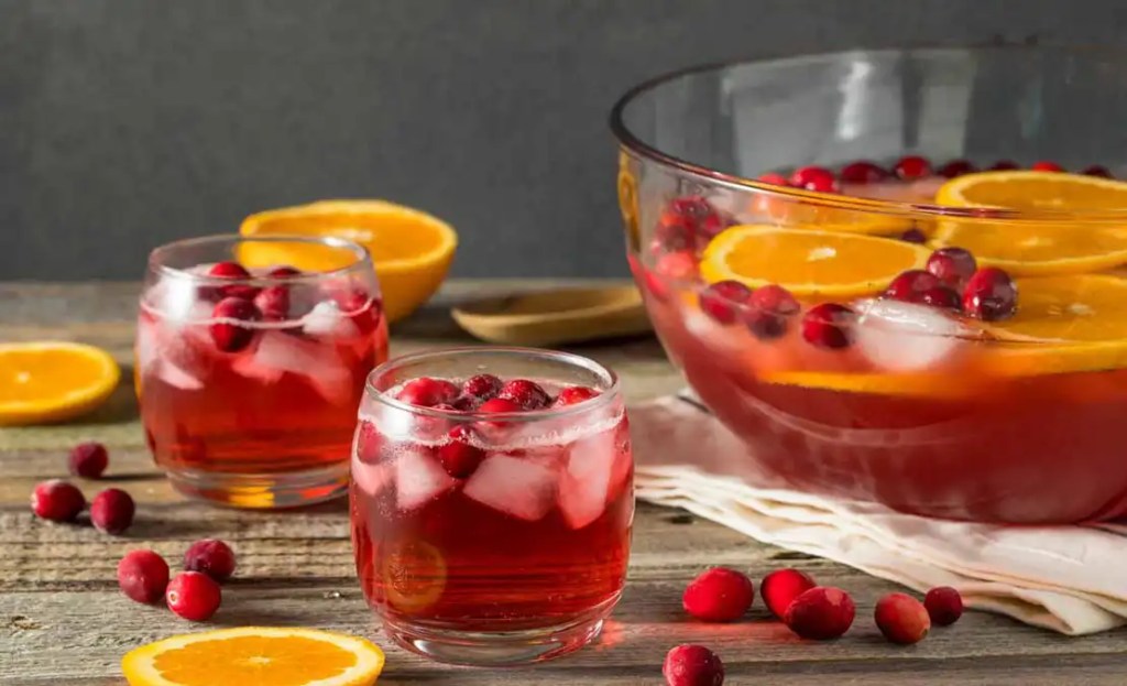 A large punch bowl filled with a vibrant cranberry-orange punch, an ideal Friendsgiving mocktail. Two glasses of the punch are garnished with fresh cranberries and orange slices, creating a refreshing and festive drink to share at a Friendsgiving gathering.
