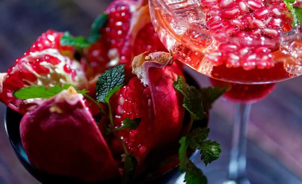 Close-up of a pomegranate cocktail garnished with fresh pomegranate seeds and mint leaves, a vibrant and unique Friendsgiving drink idea. The glass is surrounded by pomegranate halves, creating a rich, fruity presentation perfect for a festive Friendsgiving gathering.