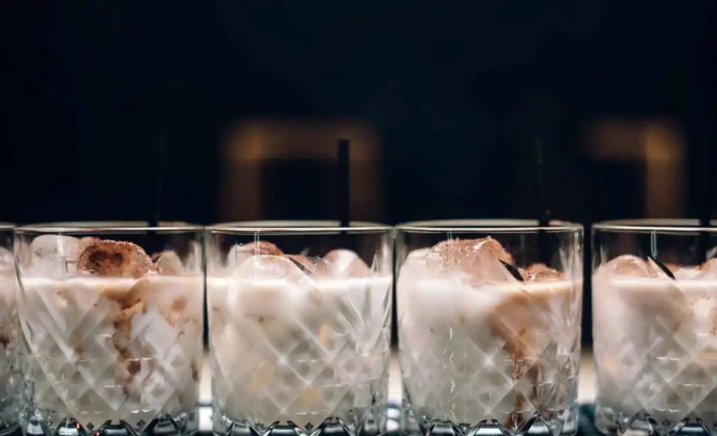 Four glasses of creamy White Russian cocktails with ice cubes, offering a rich and indulgent drink for a Friendsgiving cocktail party. The drinks are served in elegant cut-glass tumblers, creating a warm and cozy vibe perfect for a Friendsgiving celebration.