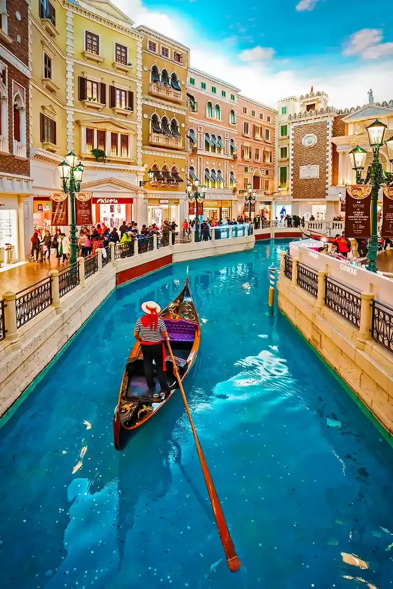 Indoor canal lined with pastel Venetian style buildings and shopfronts, with a gondolier in a striped shirt rowing a black gondola through bright blue water. Crowds stand along both sides of the walkway under decorative streetlamps, giving the scene the feel of a lively indoor shopping destination designed to resemble Venice.