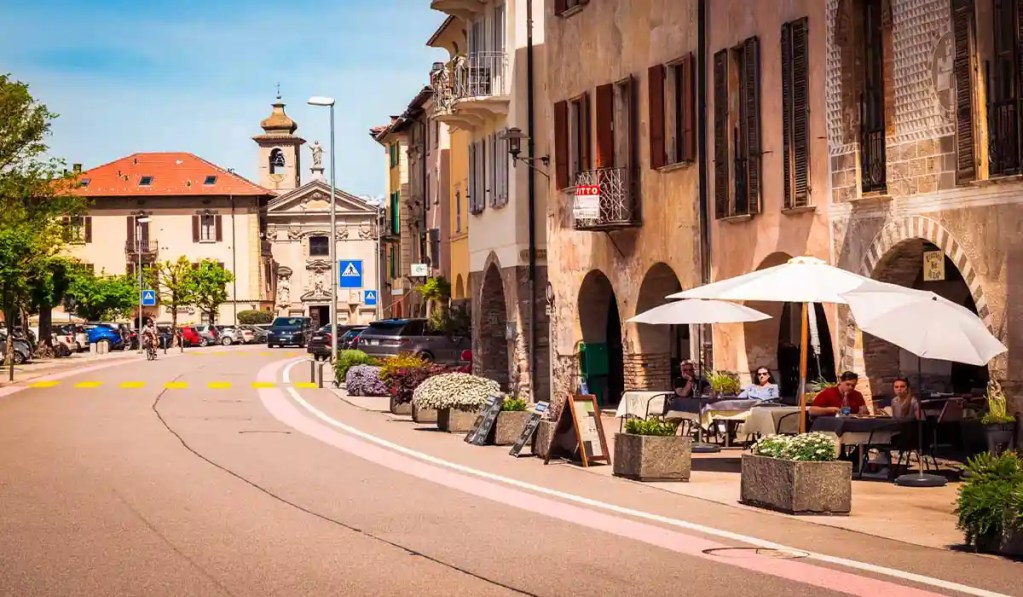 A charming street scene in Bissone, Switzerland, featuring outdoor café seating with white umbrellas along a quaint cobblestone road lined with historic buildings and colorful flowers. A church with a distinctive bell tower is visible in the background, adding to the village's picturesque atmosphere.